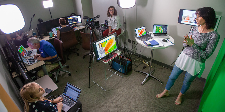 Multiple researchers working in a small room. One woman delivers a mock broadcast in front of a green screen.